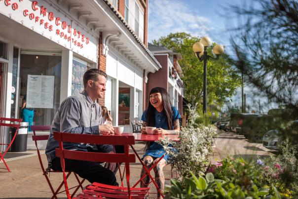 A couple sits at a small red table outside Condor Chocolates in Athens, GA, enjoying drinks surrounded by flowering plants and a vibrant street scene.