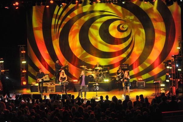 The B-52s perform live at The Classic Center in Athens, with bold yellow-orange striped backdrop seen from the darkened audience below.