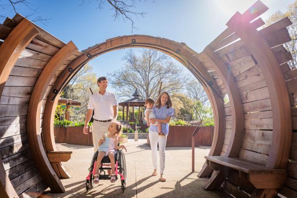 Family with children, including a girl in a pink wheelchair, walking through a wooden archway at the Children’s Garden outdoors.